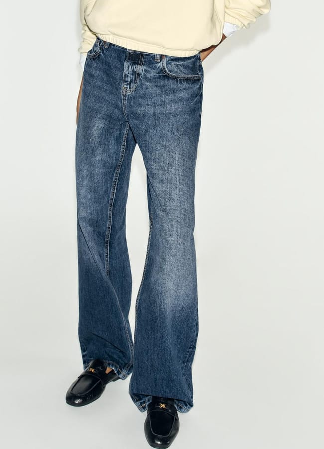 Person wearing blue flared jeans, a light long-sleeved shirt, and black shoes—an ensemble that highlights style essentials—standing against a plain white background. The person’s upper body and face are not visible.