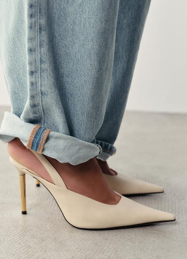 Person wearing light-wash rolled-up jeans and cream-coloured pointed-toe slingback sexy heels with gold heels, standing on a light-coloured floor.