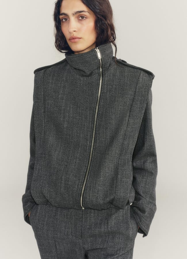 Person wearing a grey zip-up blazer with high collar and matching trousers, styled like one of this season’s standout lightweight jackets, standing with hands in pockets against a plain light background.