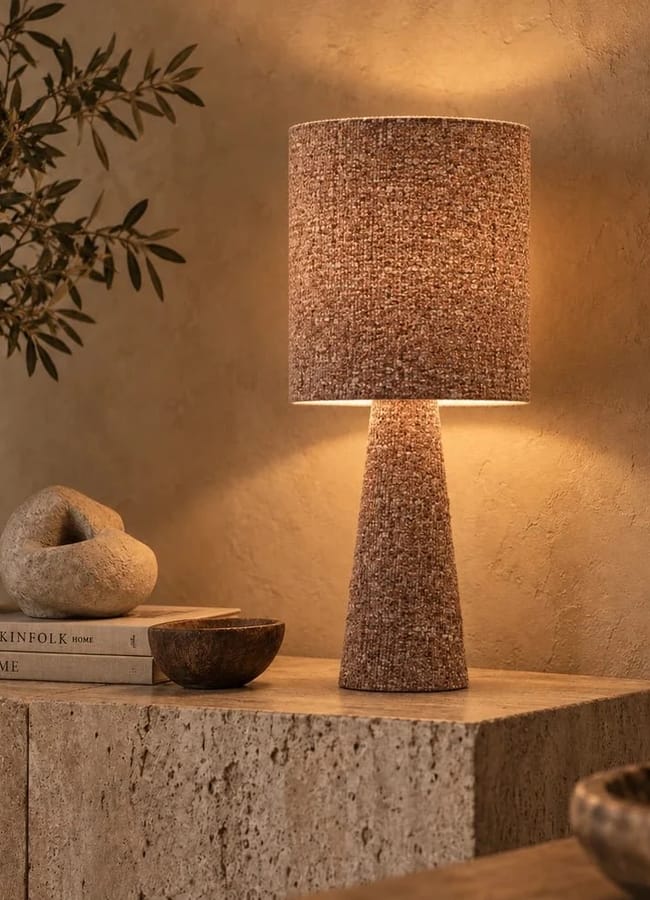 A textured bouclé cylindrical table lamp glowing on a stone console table next to a decorative stone sculpture and two stacked books.
