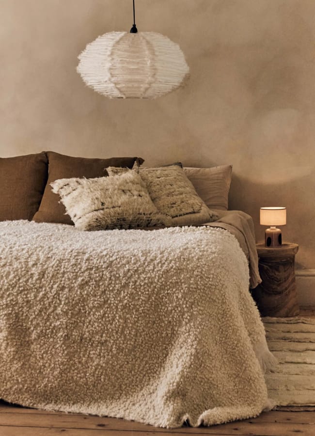 A bedroom with a bed featuring a textured white bedspread, several pillows, a pendant light, and a small bedside table with a lamp.