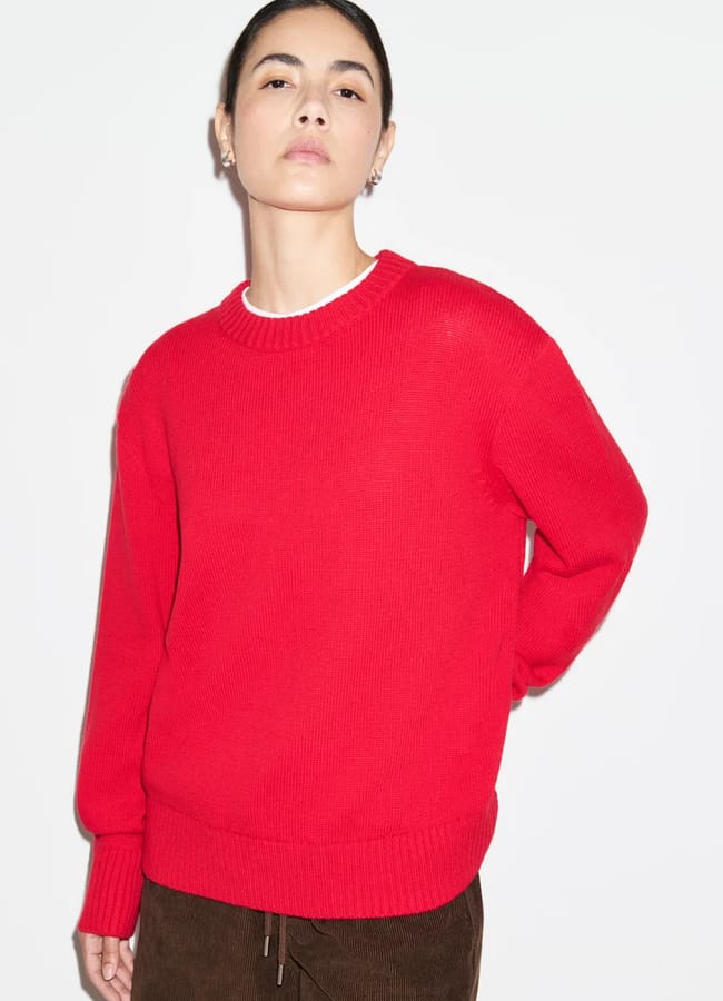 A person wearing a bright red jumper over a white shirt and brown trousers.