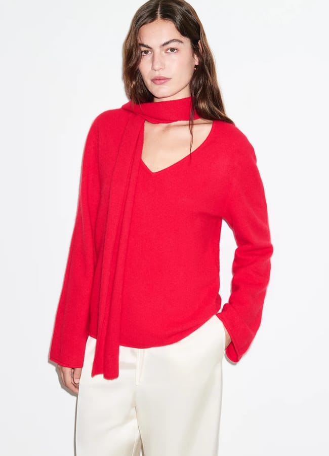 A person wearing a bright red V-neck jumper with an integrated scarf detail and cream-coloured trousers.