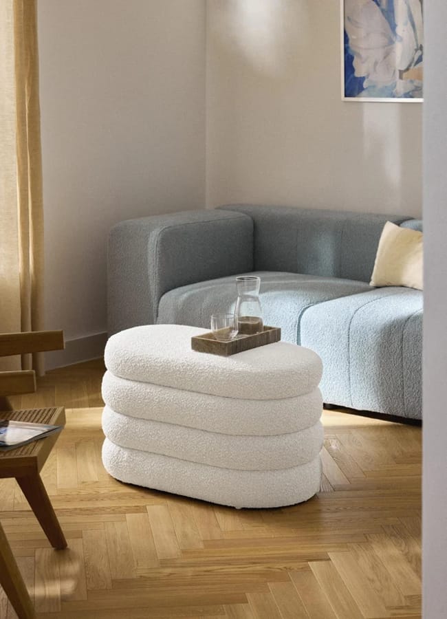 An oval, white, textured bouclé ottoman with tiered detailing, topped with a wooden tray, a glass carafe, and a tumbler.