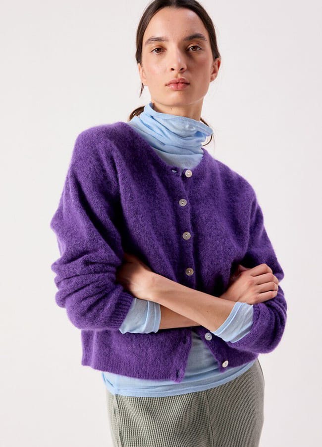 A person wearing a purple button-down cardigan over a light blue polo neck and grey trousers.