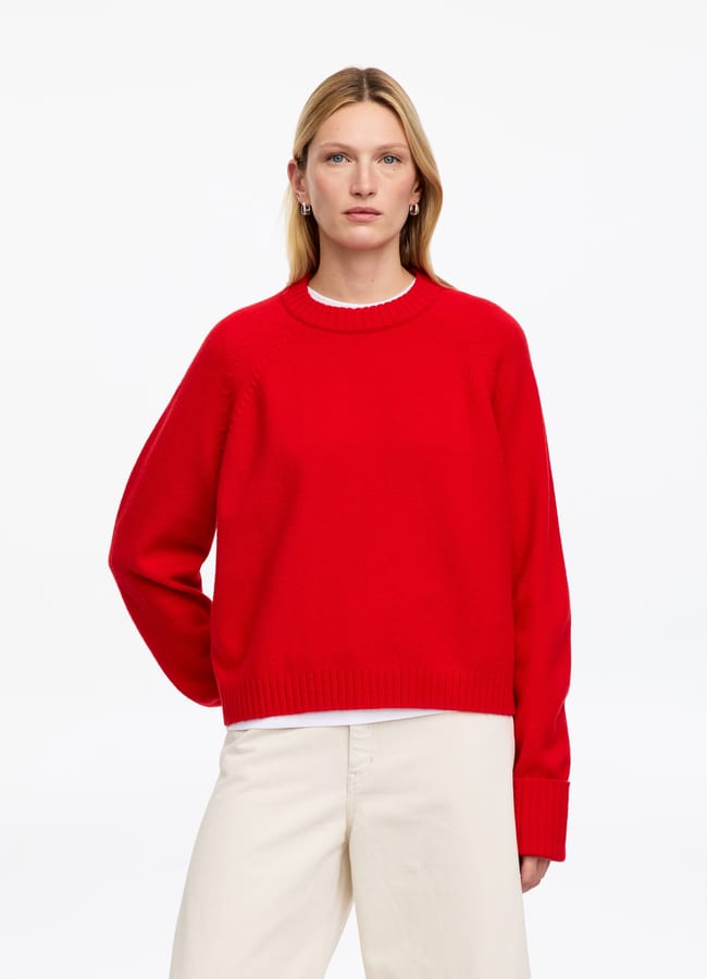 A person wearing a bright red crewneck jumper over a white shirt and cream-coloured trousers.