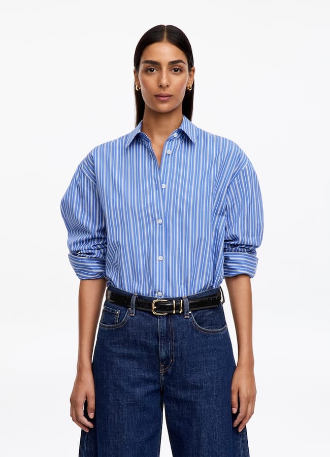 A woman wearing a blue and white striped button-down shirt tucked into dark wash jeans with a black belt.