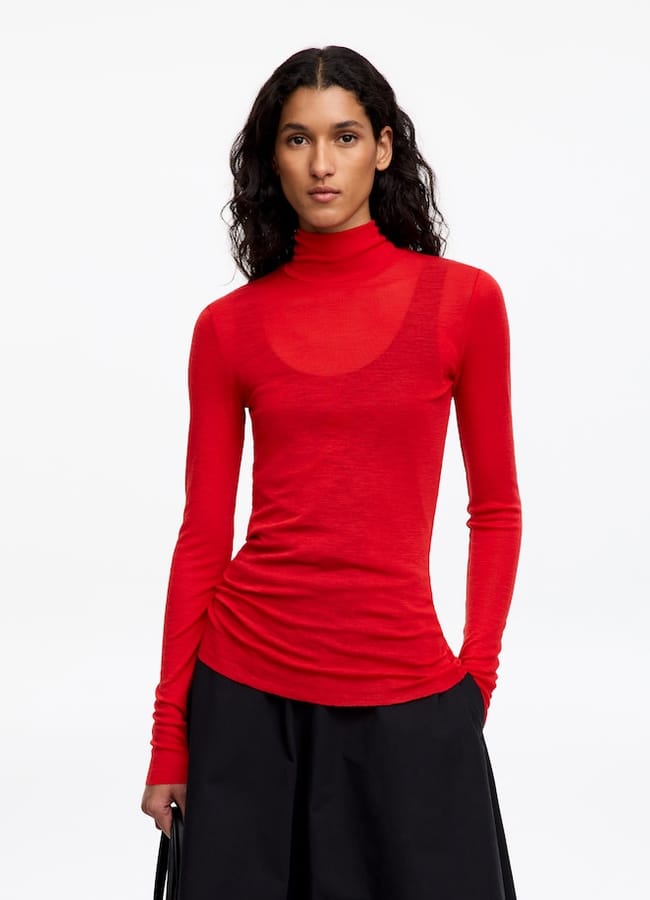 A person wearing a sheer, long-sleeved red polo-neck top over a camisole, paired with a black skirt.