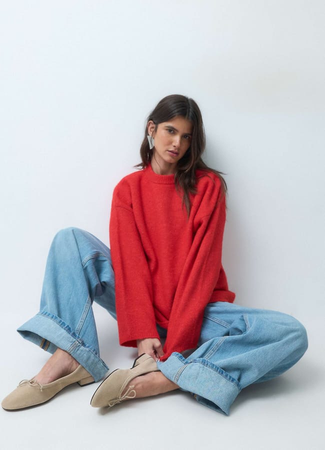 A woman wearing a red crew-neck jumper, wide-leg cuffed jeans, and tan ballet pumps sits on the floor.