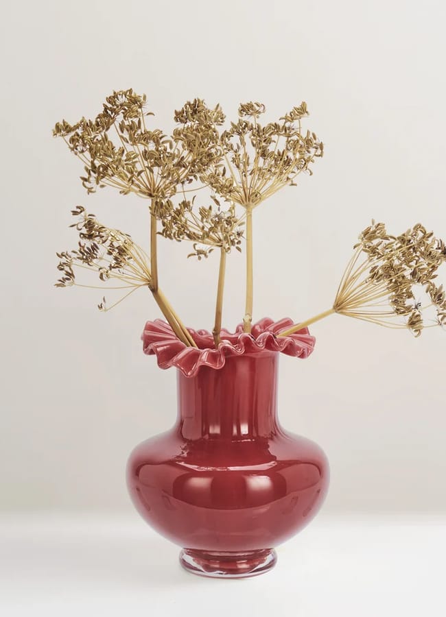 A red glass vase with a ruffled rim containing three dried botanical stems, perfect for autumn tablescape ideas.