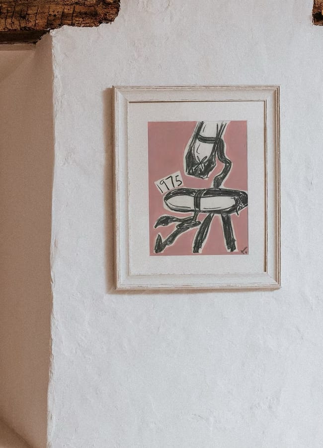 A framed line drawing of a ballet shoe on a pink background, featuring the number 1975 on a small label, from a collection of affordable female artist wall art.
