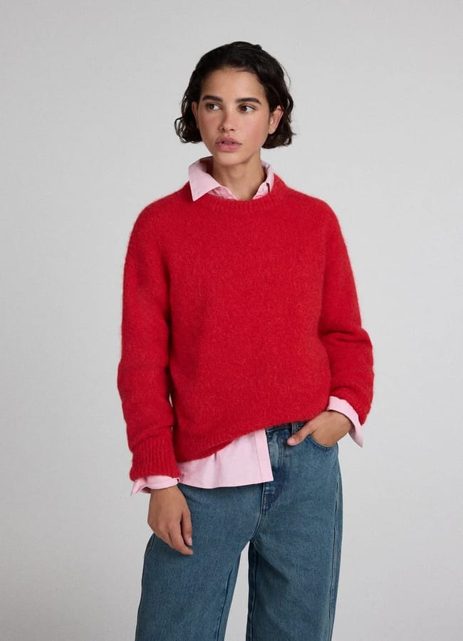 A woman wearing a fuzzy red crew-neck jumper over a pale pink collared shirt and blue jeans.