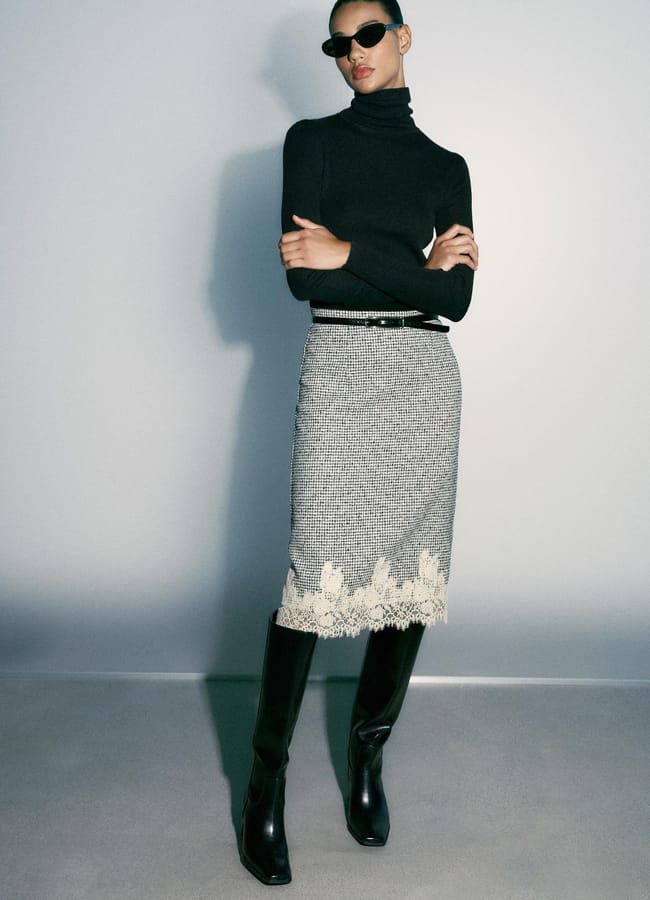 A model wearing a black polo neck, a houndstooth midi pencil skirt with a lace hem, a thin black belt, and black knee-high boots, accessorised with dark sunglasses.