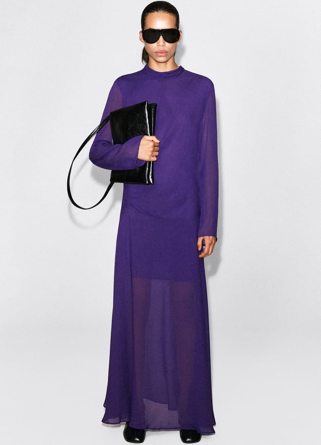 A person wearing a long-sleeved purple sheer maxi dress, black sunglasses, and black shoes, carrying a black shoulder bag.