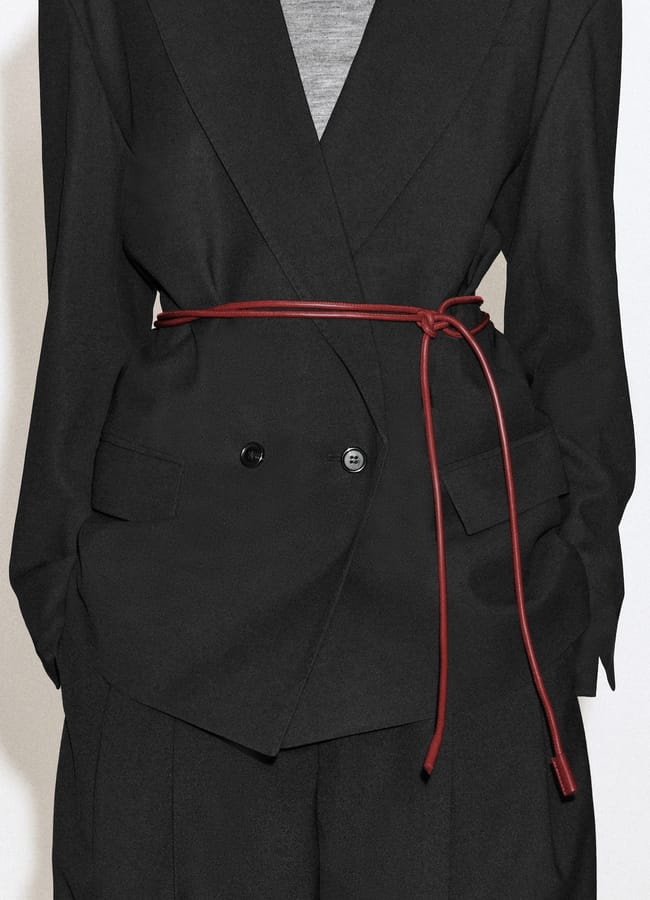 A double-breasted black blazer cinched at the waist with a thin, knotted red leather belt over a grey top.