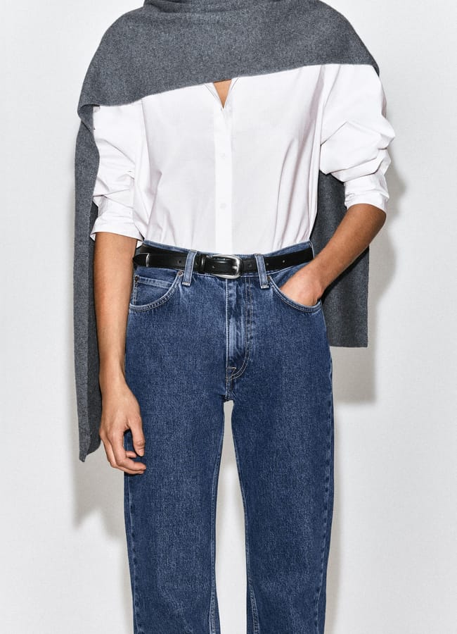 A person wearing a white button-down shirt, blue jeans with a black belt, and a grey jumper draped over their shoulders.
