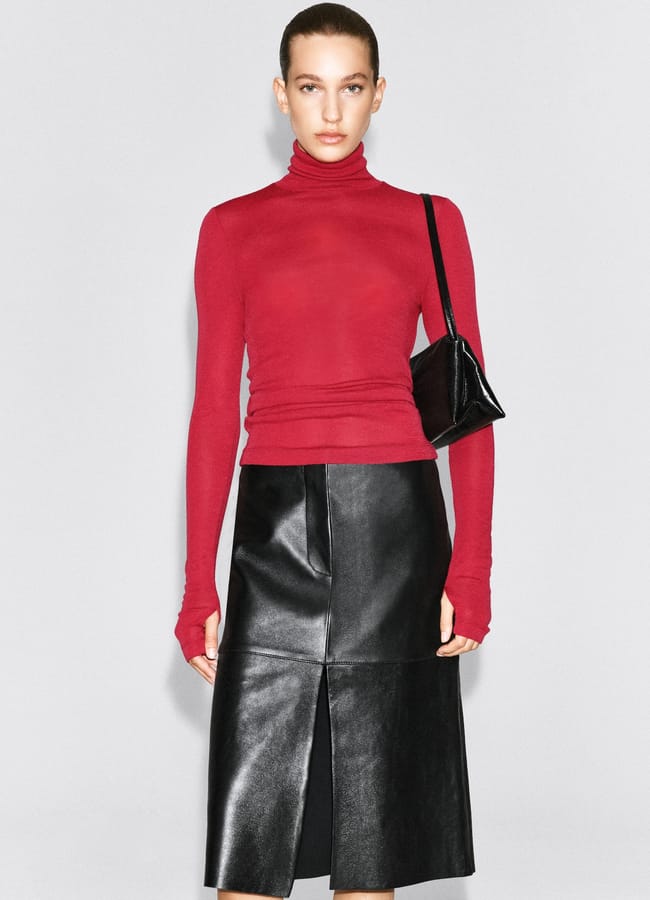 A model wears a long-sleeved red polo-neck top and a black leather skirt with a front slit, carrying a small black shoulder bag.