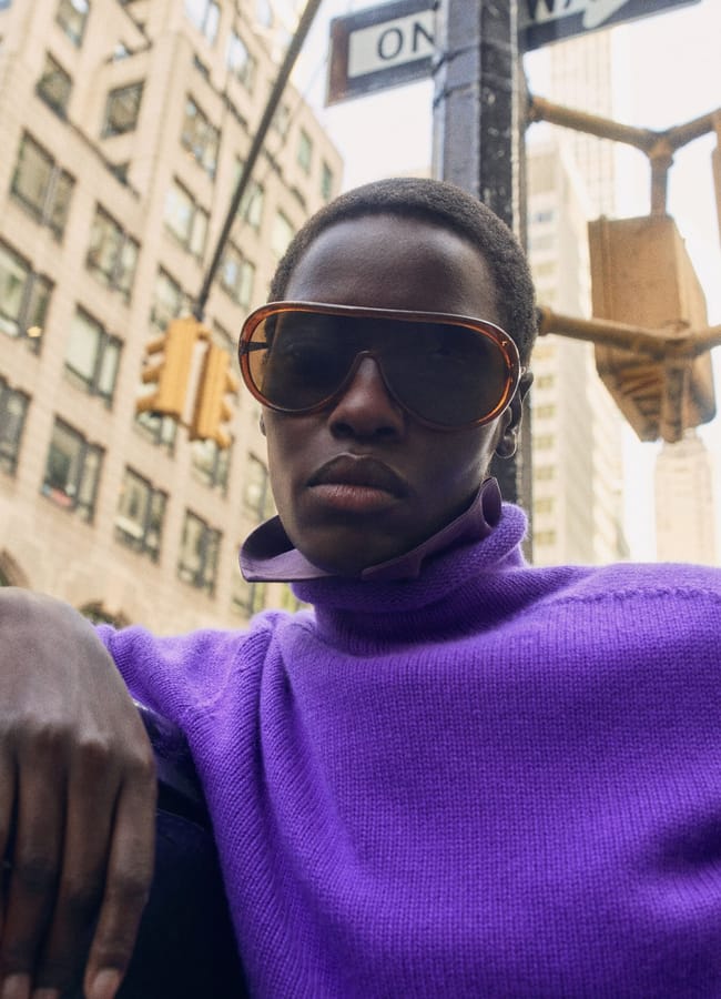 A person wearing large brown sunglasses and a purple polo neck jumper stands on a city street near a One Way sign.