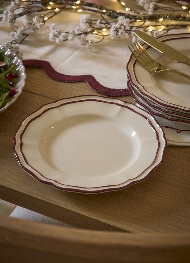 A stack of cream-coloured plates with scalloped edges and dark red rims resting on a wooden table with a fork and knife, perfect for autumn tablescape ideas.
