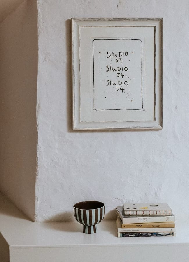A framed Studio 54 print hangs above a striped bowl and a stack of books, serving as stylish wall art on a white surface.