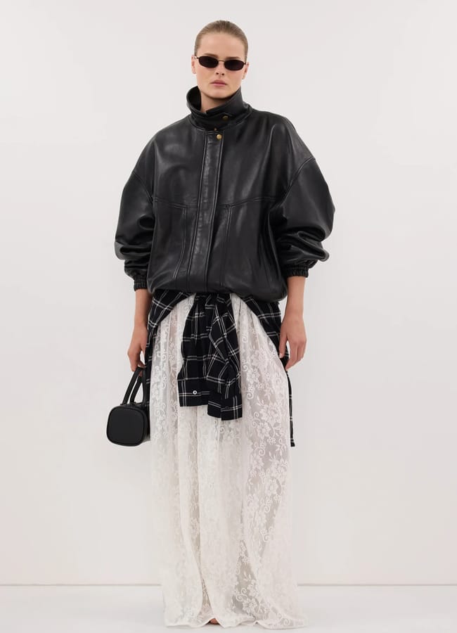 A model wearing one of the best women's leather jackets styled with a checked shirt tied around the waist, a long white lace skirt, and a small black handbag.