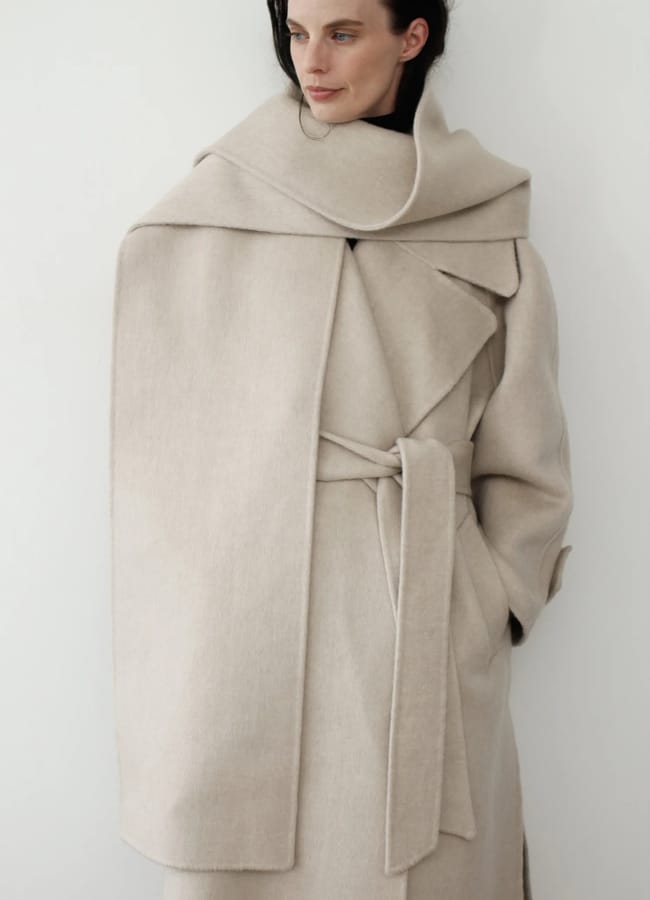 A woman wearing a belted wool coat from a black-owned fashion brand, featuring a large, draped collar in beige.