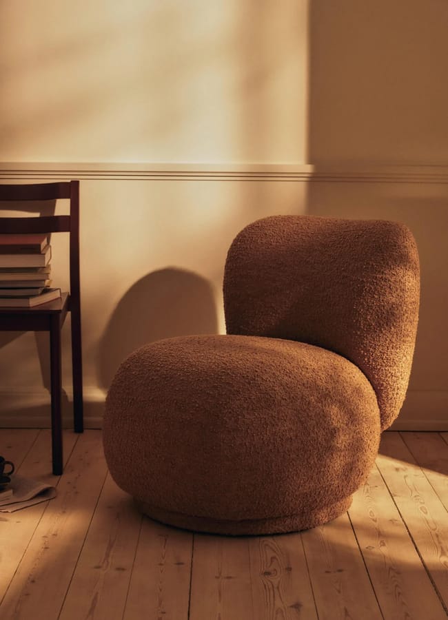 A rounded, textured brown bouclé armchair next to a wooden chair stacked with books.