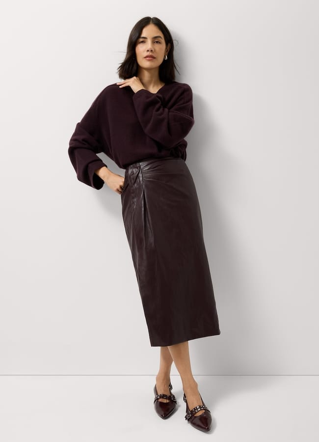A woman wearing a dark purple jumper, a dark pencil skirt with front wrap detail, and buckled pointed-toe flats.