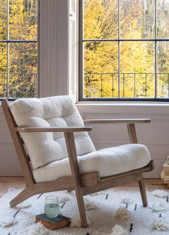A wooden armchair with white tufted bouclé upholstery sits on a patterned rug in front of large windows looking out at autumn trees.
