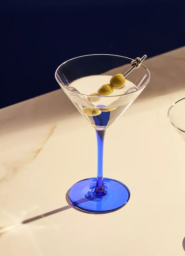 A cocktail with three olives in a blue-stemmed martini glass next to an empty matching glass on a white surface, perfect for listening room interior ideas.