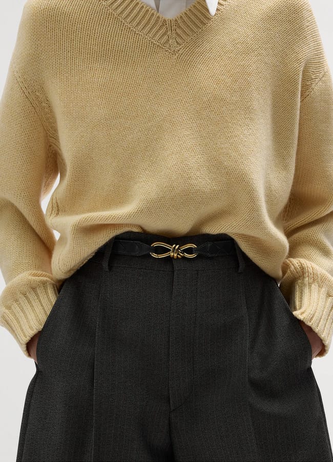 A person wearing a beige v-neck jumper over a white collared shirt, paired with grey trousers and a black belt with a gold-tone knot buckle.