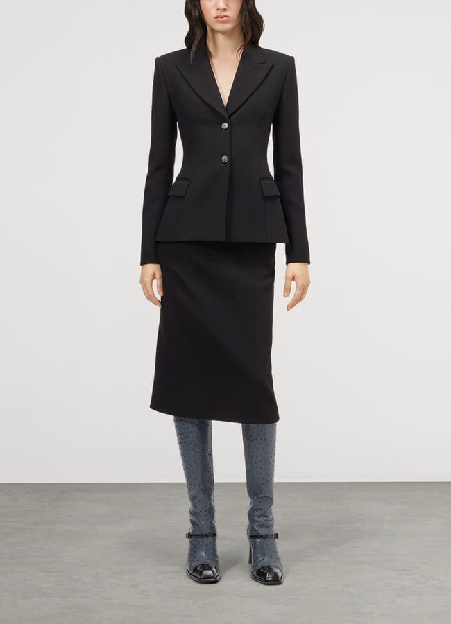 A model wears a fitted black blazer and a knee-length pencil skirt, paired with textured grey knee-high boots.