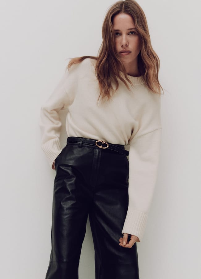 A woman wearing a cream-coloured jumper tucked into black trousers with a gold-buckle belt.