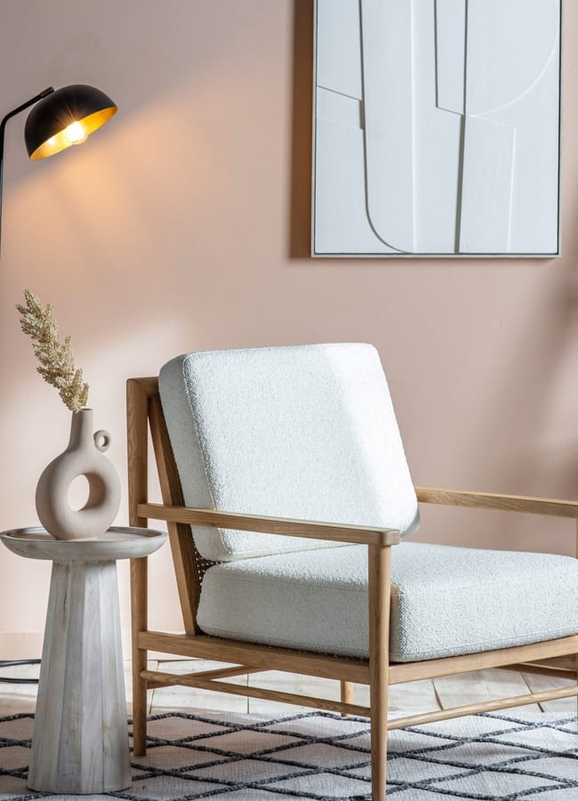 A light wood armchair with a white bouclé cushion sits next to a small pedestal table holding a ceramic vase with dried stems.