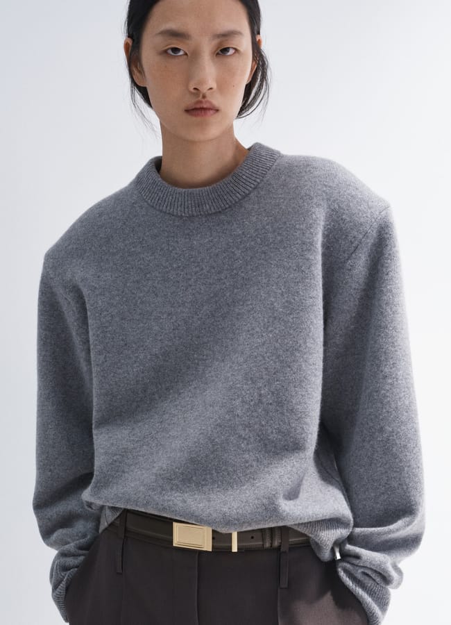 A person wearing a grey crew-neck jumper tucked into dark trousers with a gold-buckled belt.
