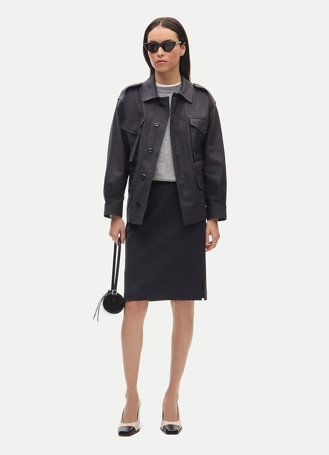 A woman wears a dark utility jacket, grey jumper, and black pencil skirt, carrying a small circular bag and wearing cat-eye sunglasses and two-tone heels.