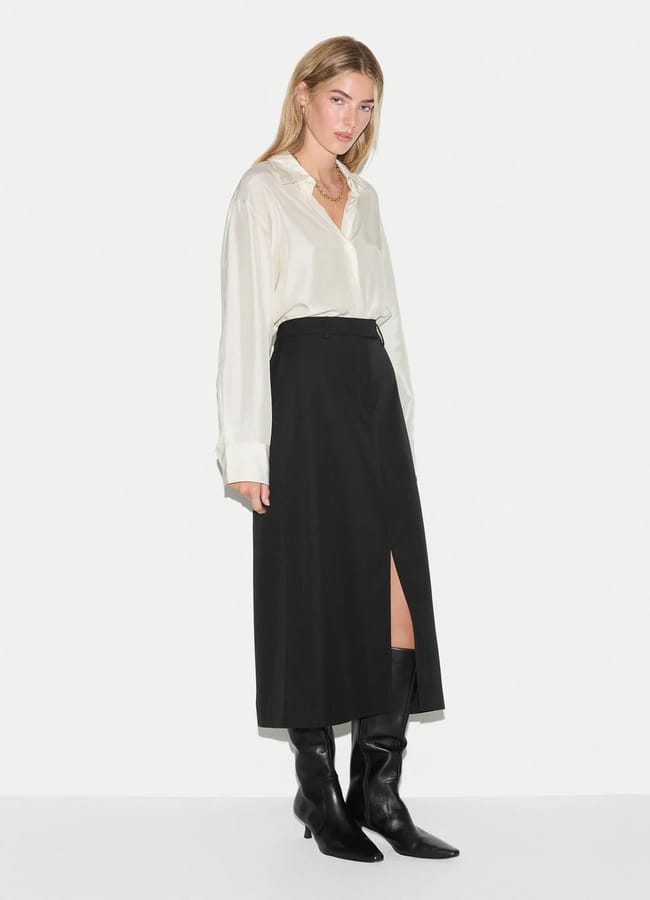 A woman wearing a cream button-through shirt, a black midi pencil skirt with a front slit, and black knee-high boots.