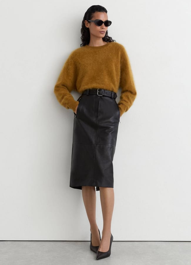 A person wearing a fuzzy mustard-yellow jumper, a black pencil skirt with a belt, and black court shoes.