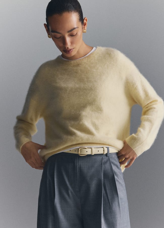 A person wearing a pale yellow crew-neck jumper, grey trousers, and a thin belt.