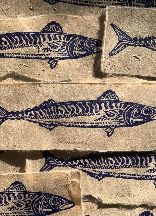 Multiple rectangular scraps of light-coloured fabric featuring affordable wall art, each printed with a dark blue linocut design of a mackerel fish.