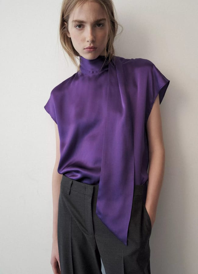 A person wears a sleeveless, high-neck purple blouse with a scarf-style tie front and grey trousers.