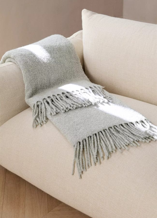 A sage green bouclé tasseled throw draped over a beige sofa.