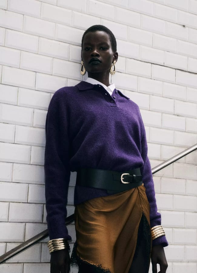 A person wearing a purple long-sleeved polo jumper, a wide black leather belt, and a brown skirt, leaning against a white tiled wall.