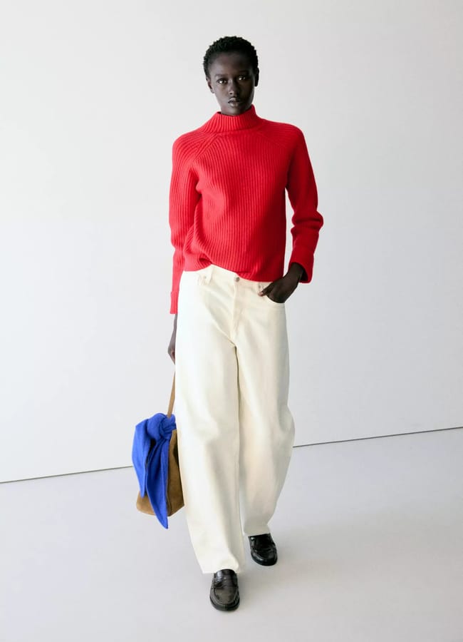 A person wears a red ribbed polo-neck jumper and cream-coloured wide-leg trousers, carrying a tan bag with a bright blue scarf attached.