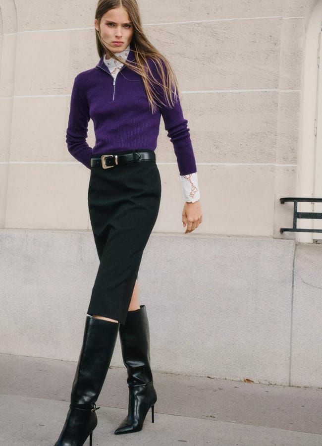 A woman wearing a purple quarter-zip jumper over a lace-collared blouse, a black pencil skirt with a belt, and black knee-high heeled boots.