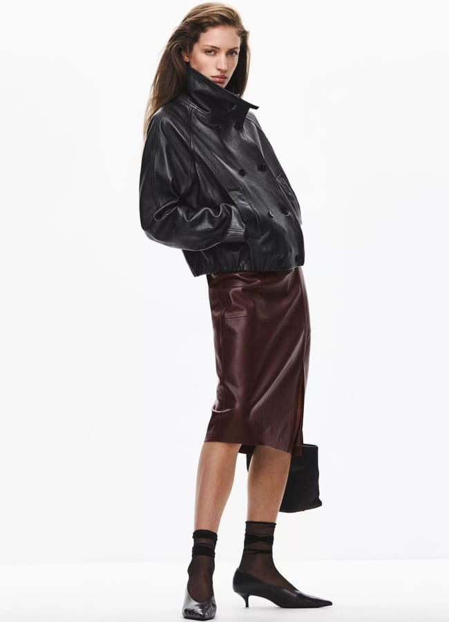 A woman poses in a black leather jacket, a burgundy midi pencil skirt, sheer black tights, and pointed-toe heels whilst holding a black handbag.