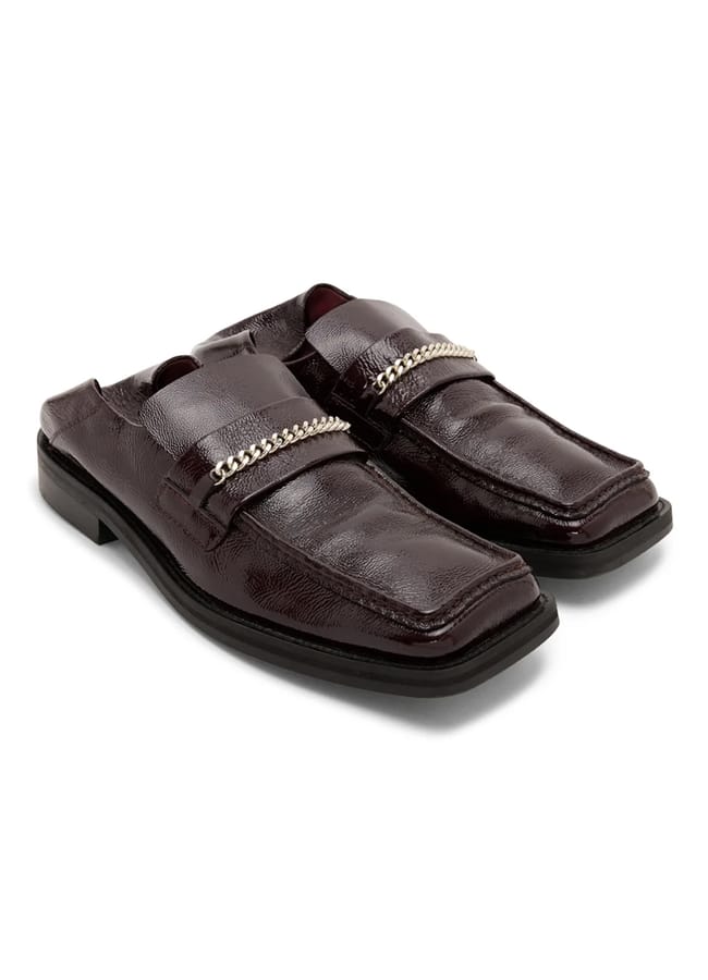 Dark brown square-toe mule loafers from a black-owned fashion brand, featuring a silver chain detail across the front.