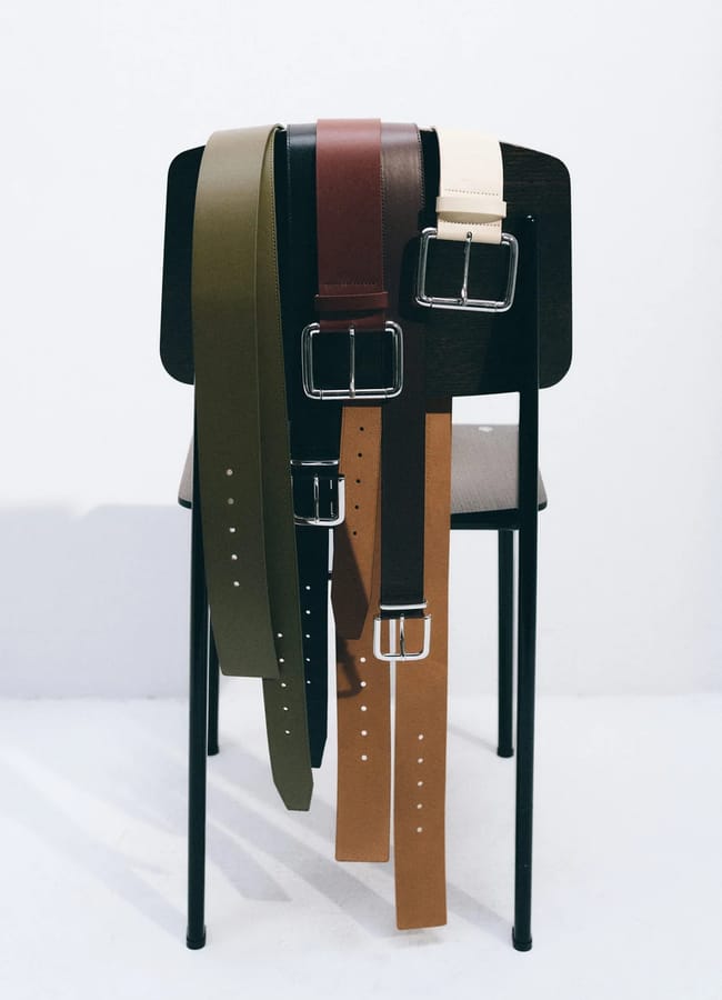 Four leather belts in olive, black, burgundy, and tan draped over the back of a wooden chair.