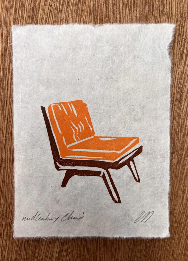 A linocut wall art print of a mid-century style chair with an orange seat and dark brown frame on textured white paper.