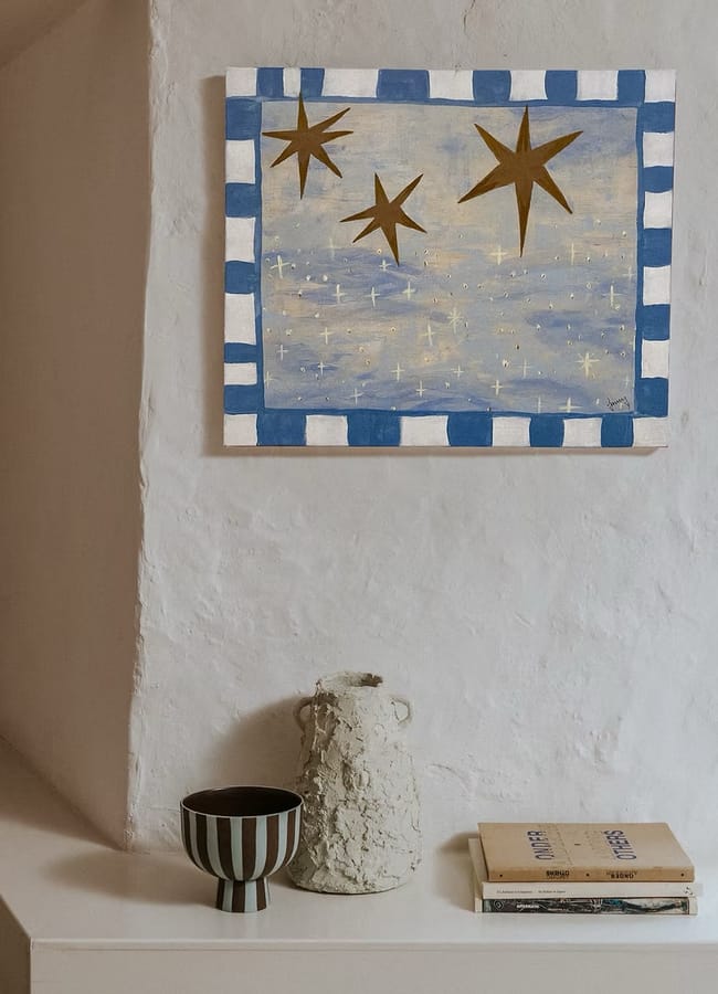 A painting of three large stars above a field of smaller stars, framed by a blue and white chequered border, hangs on a white wall as affordable wall art above a textured vase, a striped bowl, and a small stack of books.